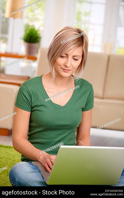Blond woman sitting on floor at home in living room using laptop computer for browsing internet