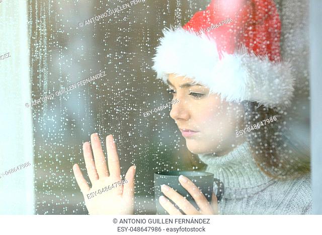 Sad and lonely woman looking through a window touching glass, with the hand in christmas time in a rainy day