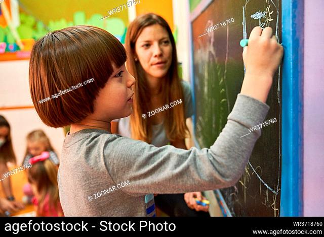 Junge beim malen mit Kreide an der Tafel in der Vorschule oder im Kindergarten mit Erzieherin