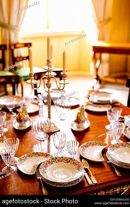 Festive table setting with clean empty plates, spoons, knives, wine glasses, decanters and a candlestick in the center on a wooden table in the room