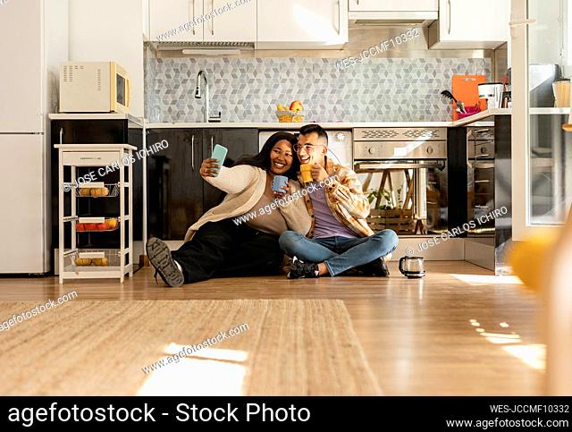 Happy couple taking selfie holding coffee cup in kitchen
