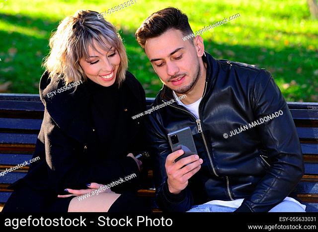 Two young friends sitting on a park bench talking consulting the mobile
