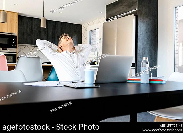 Mature businessman with hands behind head sitting at table in home office
