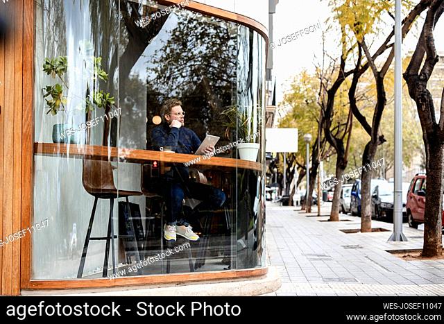 Businessman with tablet PC sitting in coffee shop seen through glass window