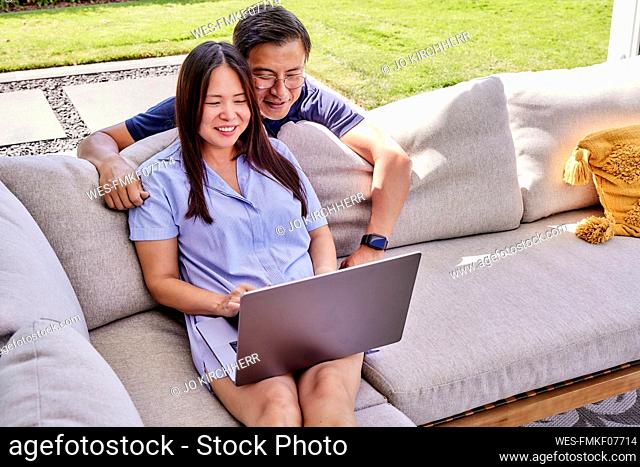 Smiling mature freelancer sharing laptop with man leaning on sofa