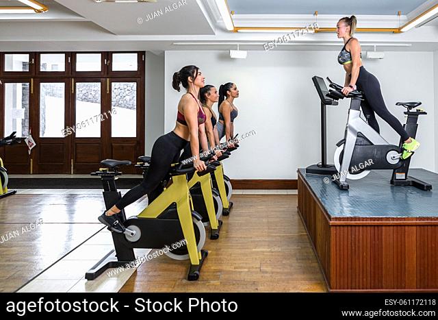 Side view of sportswomen and female instructor leaning on handlebar of exercise bike during fitness training in light gym