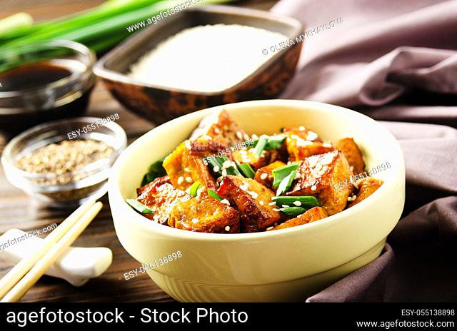 Crispy stir fried tofu cubes with chives in clay dish on wooden kitchen table with napkin chives pepper and soy sauce aside