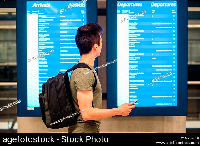 Young man standing in the railway station next to screens with schedule, planning a travel, travelling with backpack