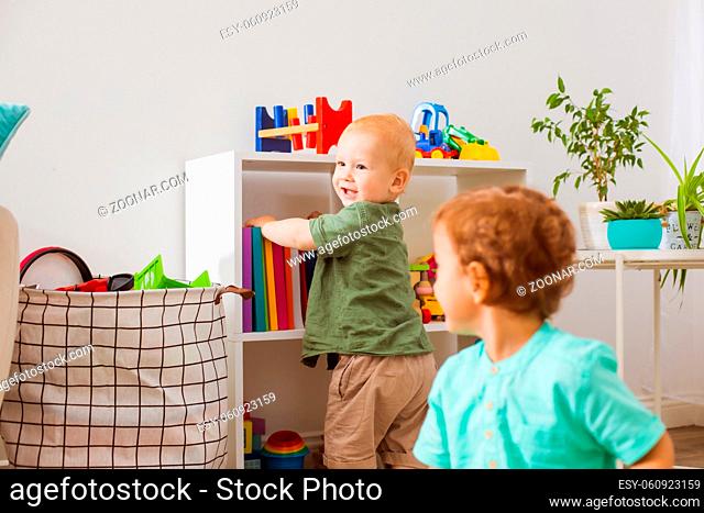 Two babies in the playroom choose toys. Focus on the boy who is standing near the bookcase in the background