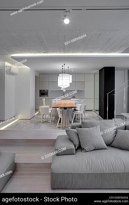 Modern interior with white walls and a stair with metal railing. There is a gray sofa with pillows, kitchen zone with wooden table and chairs