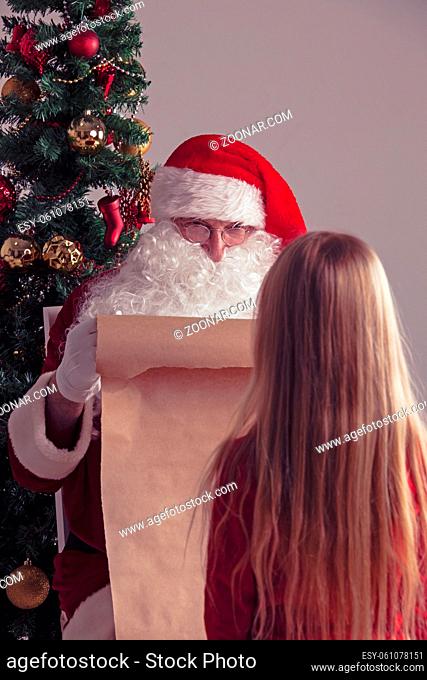 Santa Claus with with list or nice or naugthy list paper with small girl near christmas tree
