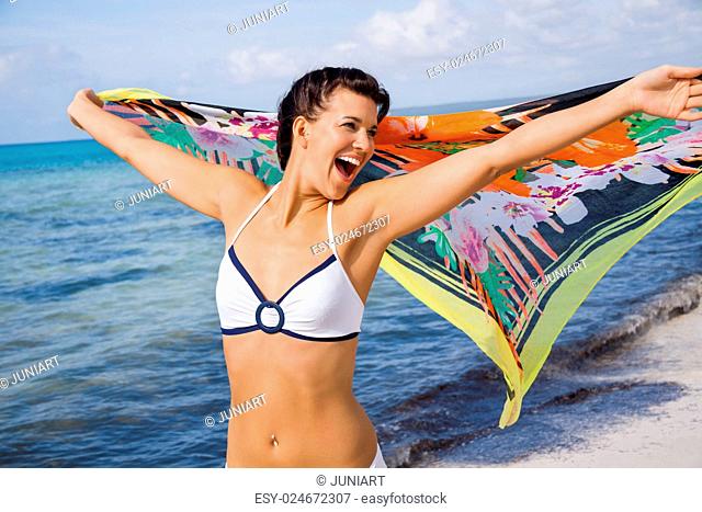 Laughing vivacious woman in a bikini at the seaside holding a colourful patterned scarf in her outstretched hands to flutter in the breeze against an ocean...