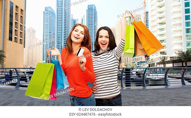 shopping, sale, tourism and people concept - two smiling teenage girls with shopping bags and credit card over dubai city skyscrapers background