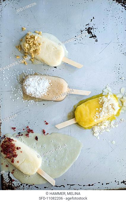 Four Ice Cream Popsicles Sprinkled with Assorted Toppings, Melting on a Metal Surface, From Above