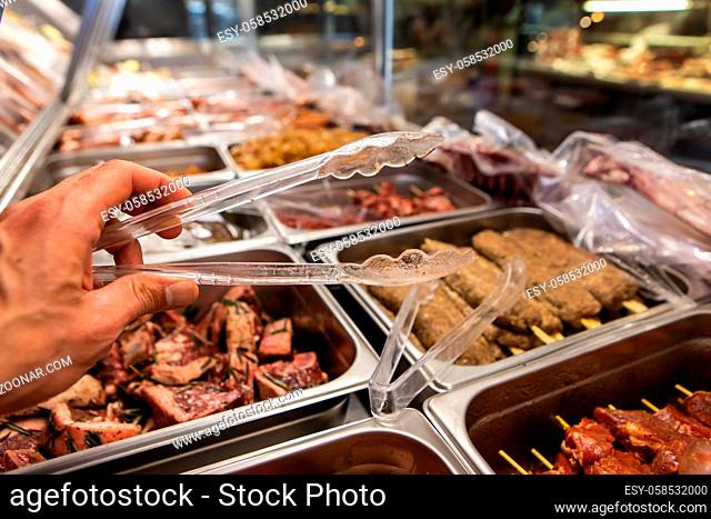 Selective focus view of butcher hand holding plastic tong with a variety of meat products ready for cooking on the display refrigerator in background