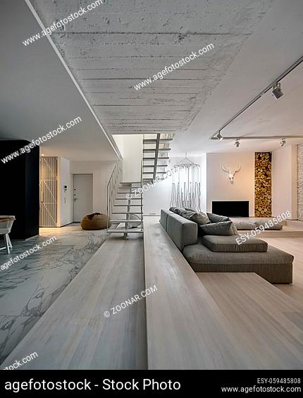 Hall in a modern style with white walls. There is a white wooden stair with a metal railing, gray sofa with pillows, glowing tube lamps, brown pouf, door