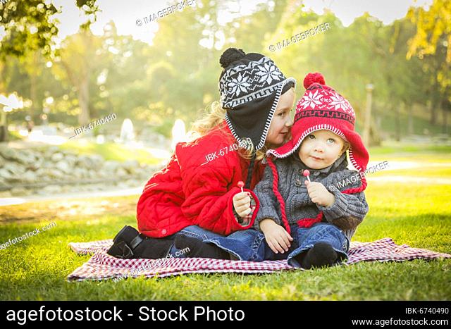 Little girl whispers A secret to her baby brother wearing winter coats and hats sitting outdoors at the park