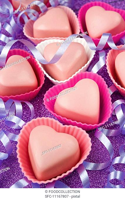 pink love heart shaped chocolates in pink cake covers with purple party streamers on a purple glittery table top
