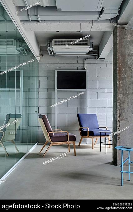 Interior in the office in a loft style with a light brick wall. There are metal round tables and multi-colored armchairs with wooden legs between a concrete...