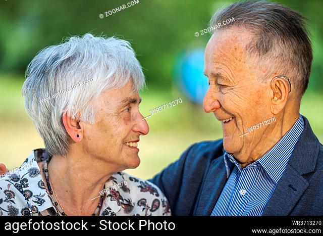 Head-and-shoulder portrait of a lovingly smiling senior couple with glasses in front of the blurred background of a park