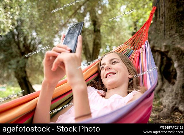 Girl lying in hammock in an olive grove, using smartphone