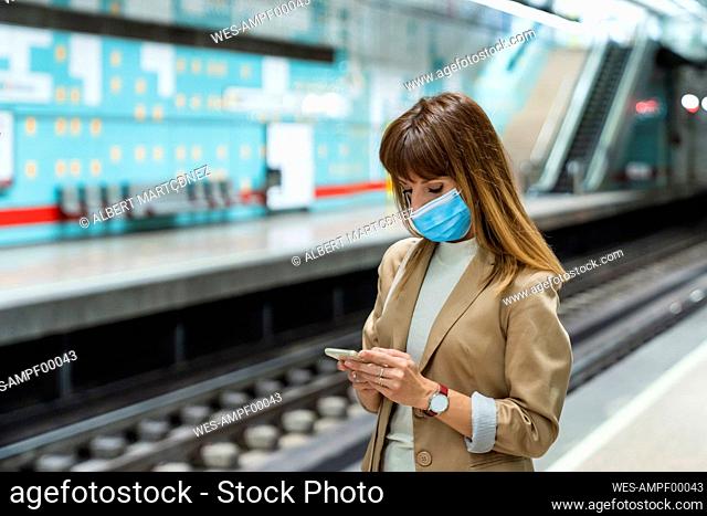 Woman with protective face mask using mobile phone while standing at subway station during pandemic