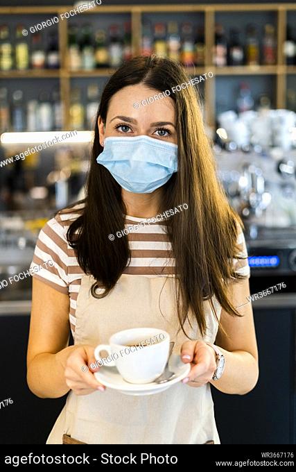 Beautiful young owner wearing mask while holding coffee cup and saucer in cafe