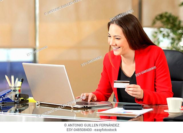 Portrait of a businesswoman wearing red suit buying on line with a credit card in a laptop sitting in a desk at office