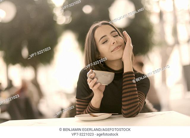 portrait of pleased woman holding coffee cup while enjoying break at table in café, closed eyes, pure joy, in Munich, Germany