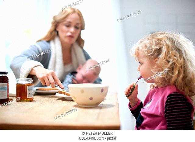 Mother holding baby boy, sitting at kitchen table with young daughter, having breakfast