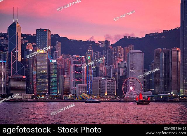 HONG KONG, CHINA - MAY 1, 2018: Tourist junk boat ferry with red sails and Hong Kong skyline cityscape downtown skyscrapers over Victoria Harbour in the evening