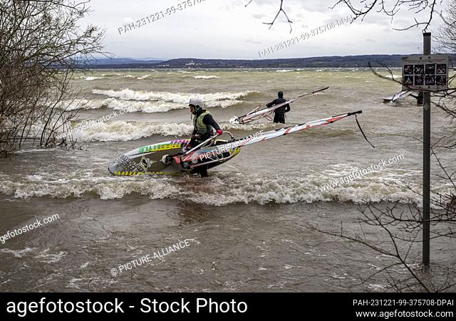 21 December 2023, Bavaria, Munich: Two surfers carry their boards into the choppy waters of the Ammersee near Herrsching in strong winds