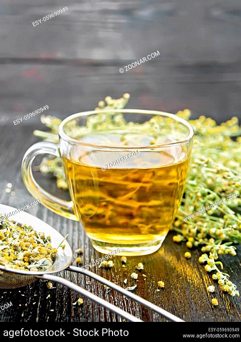 Gray wormwood herbal tea in a glass cup, fresh flowers and metal strainer with dry sagebrush flowers on wooden board background