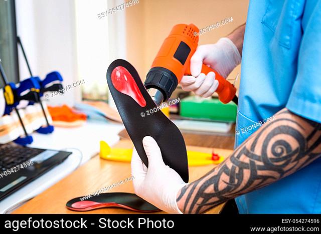 closeup Hands of young man with tattoo in workshop dressed in blue uniform make individual orthopedic insoles. The instrument uses hairdryer to heat and deform