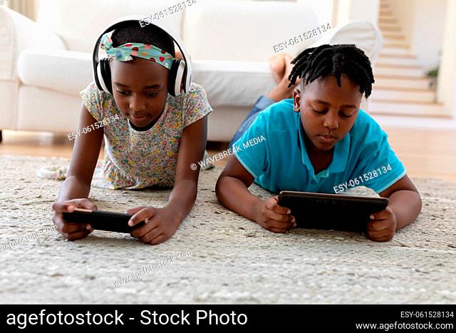 African american brother and sister wearing headphones lying on floor using smartphones