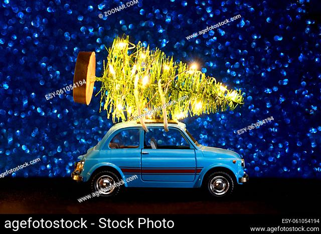 Blue retro toy car delivering Christmas or New Year illuminated tree on blue festive background