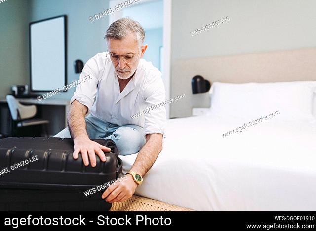 Man closing suitcase while sitting on bed in hotel room