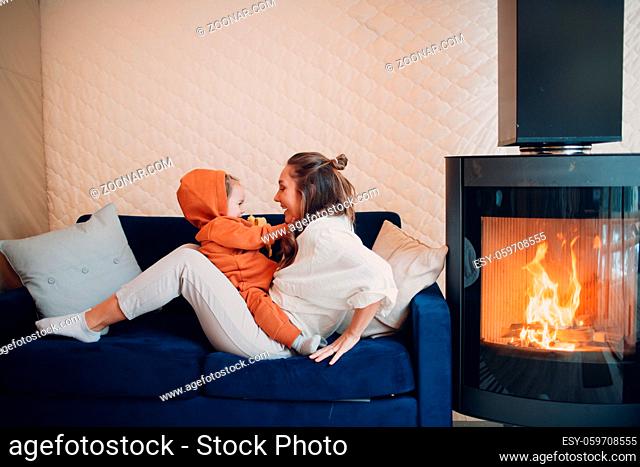Mother and child sitting and playing on sofa near fireplace. Single parenting. Mom and baby child. Parent and little kid relaxing at home