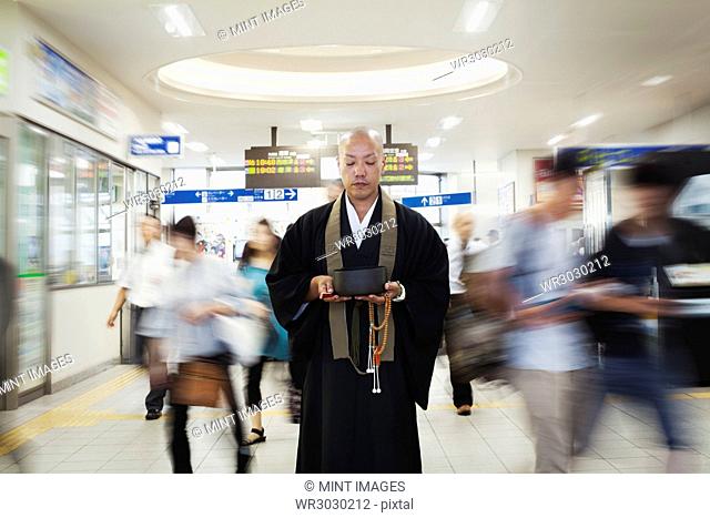 Buddhist monk with shaved head wearing black robe standing inside a train station, holding singing bowl and mala, people walking past