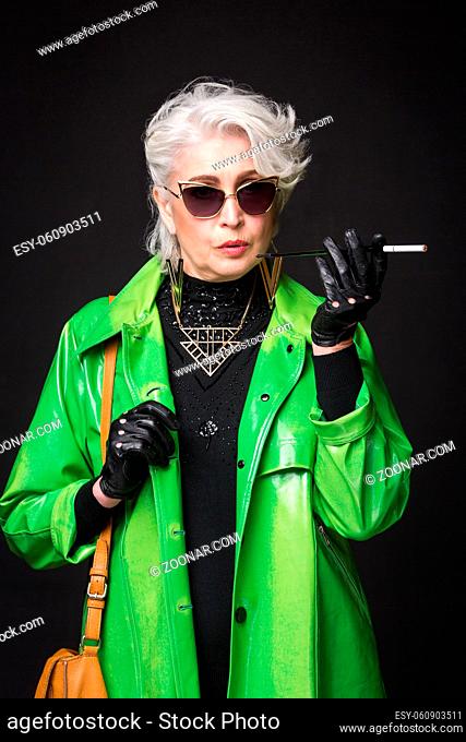 Portrait of senior rich woman in bright green coat wearing sunglasses while holding cigarette in front of her isolated on black background