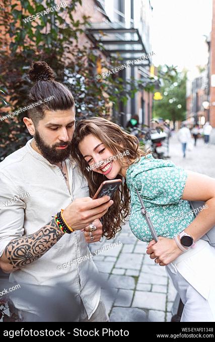 Hipster man showing smart phone to girlfriend while standing on footpath