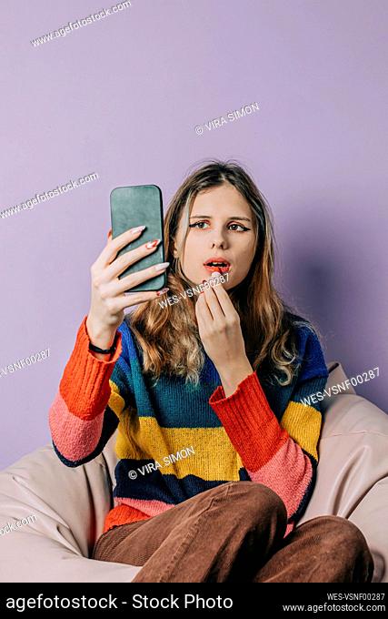 Girl looking at smart phone and applying lipstick against purple background