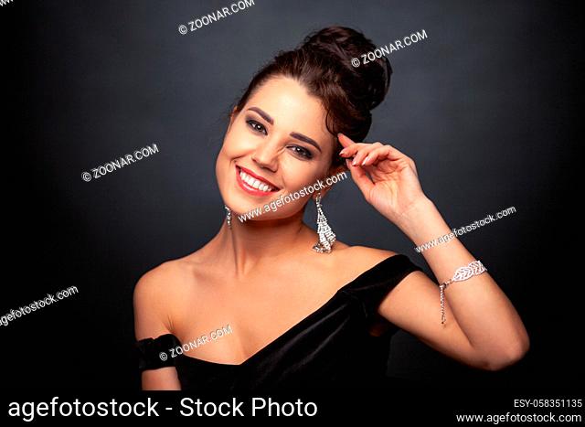 Beautiful brunette lady with hair styling in a black evening dress wears earrings and a bracelet with red gems. Studio photo on a gray background
