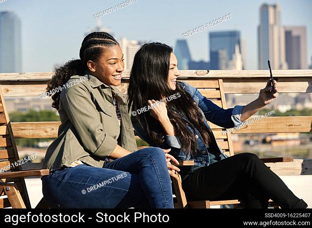 Group of young co-workers hanging out on rooftop patio, two women taking selfie with mobile phone