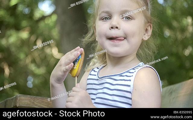 Little blonde girl holds gingerbread in Ukrainian national colors in her hand, it says ""Ukraine is me"". Close-up portrait of girl sitting on park bench with...