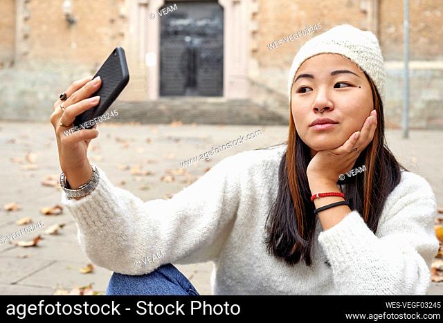 Young woman with hand on chin listening to video call while sitting on footpath