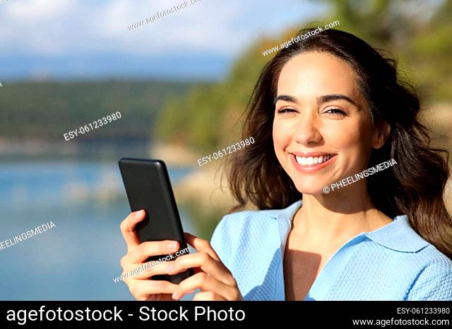 Happy woman holding smart phone looking at you in nature