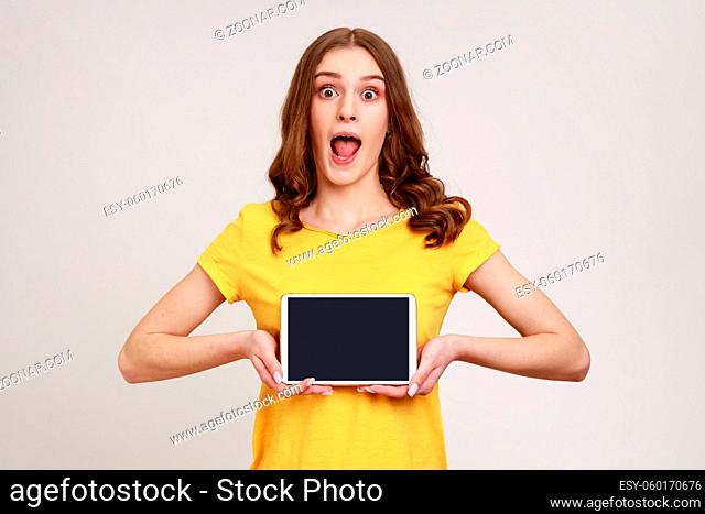 Portrait of shocked beautiful teenage girl in style T-shirt, showing tablet empty screen with unbelievable face, looking at camera with opened mouth