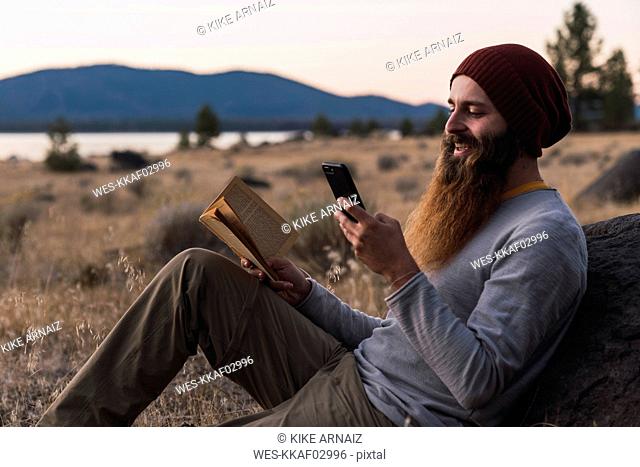 USA, North California, bearded young man with book and cell phone during a hiking trip near Lassen Volcanic National Park