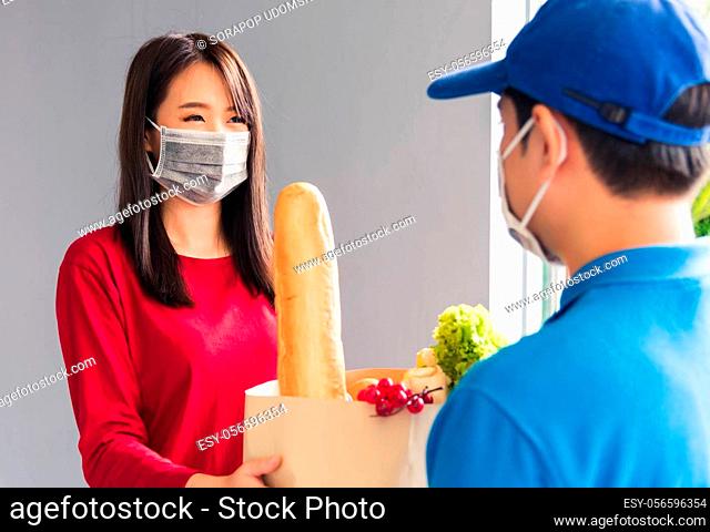 Asian young delivery man in uniform wear protective face mask he making grocery service giving fresh food to woman customer receiving front house under pandemic...